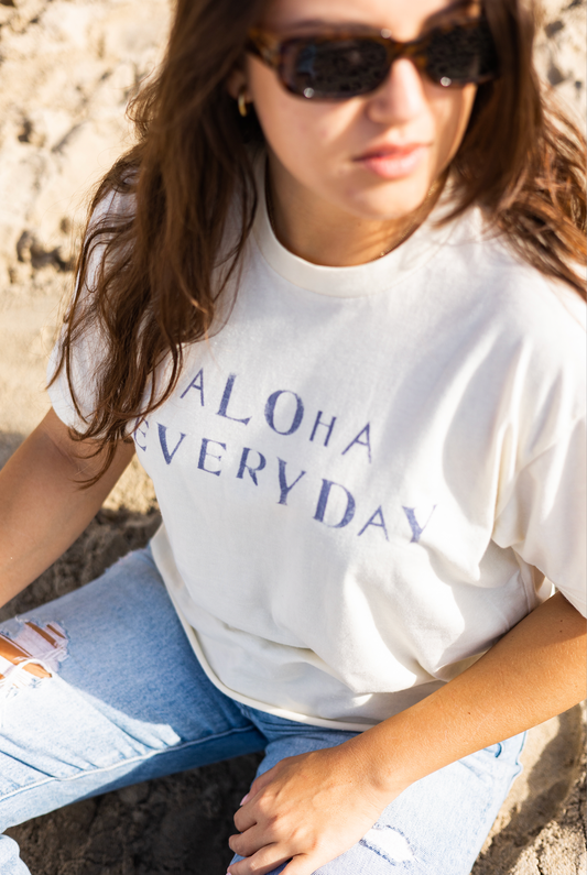 Woman wearing a 'Aloha Everyday' t-shirt sitting on a stone surface.